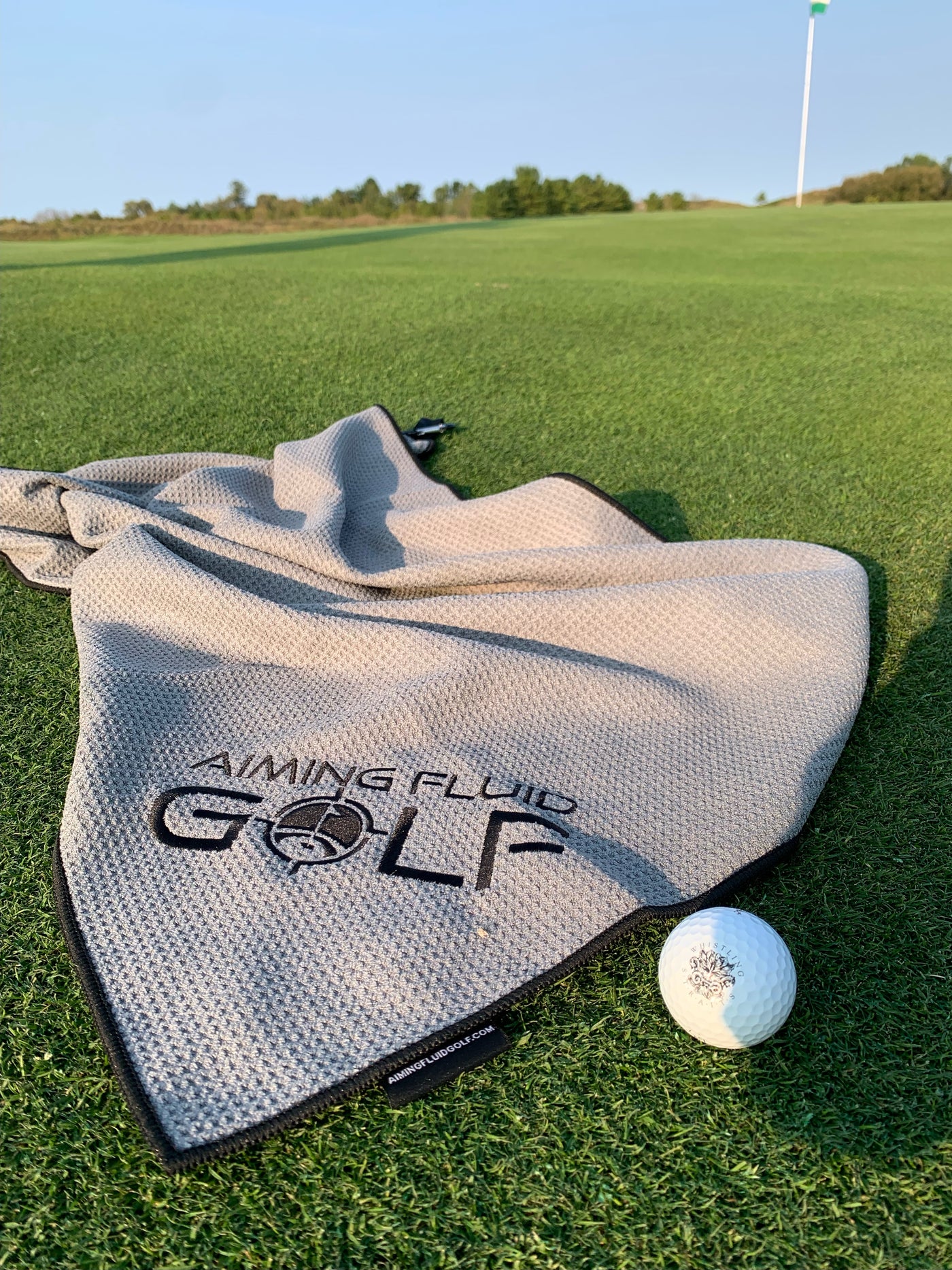 Aiming Fluid Golf (THE BEST TOWEL IN GOLF) Magnetic Towel Tall Boy (Large) in color grey laying on the green